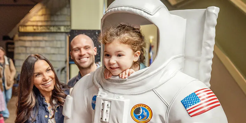 Kid poking her head out of a demo astronaut suit as parents look on smiling