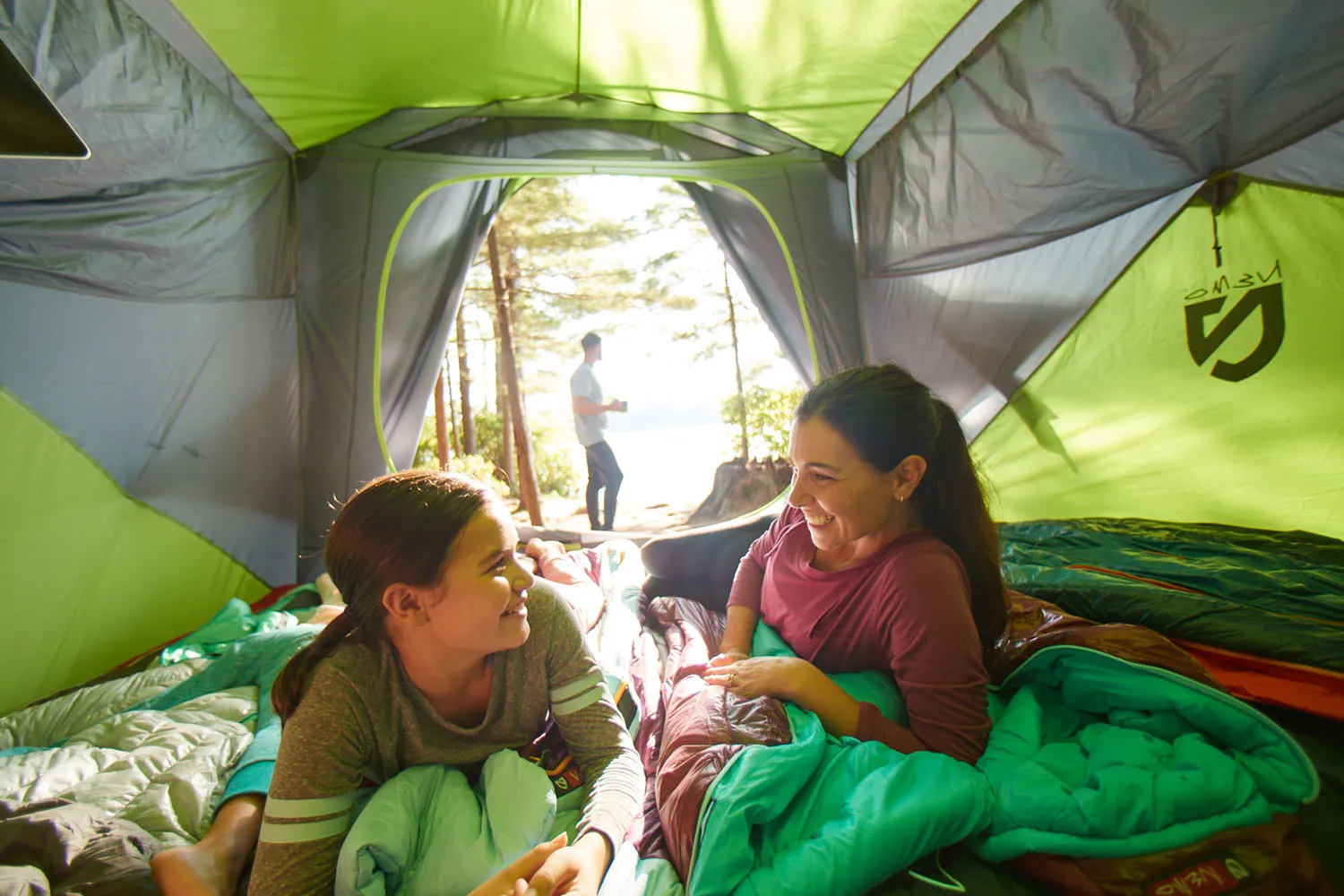 Mother and daughter smiling laying on sleeping bags in a tent with the sun shining in