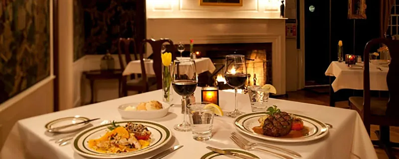 A close-up view of a set dining table with two plates of food, glasses of wine and water, and silverware. A fireplace with a lit fire is in the background