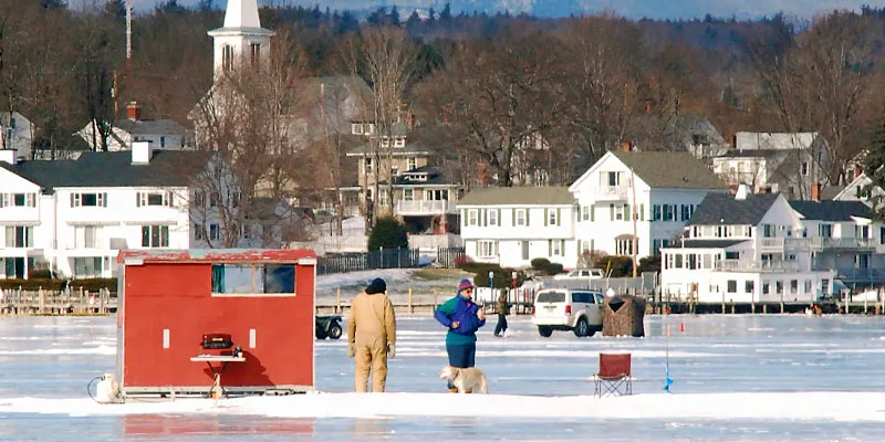 People stading outside of a bobhouse on a frozen lake with a small New England town in the background