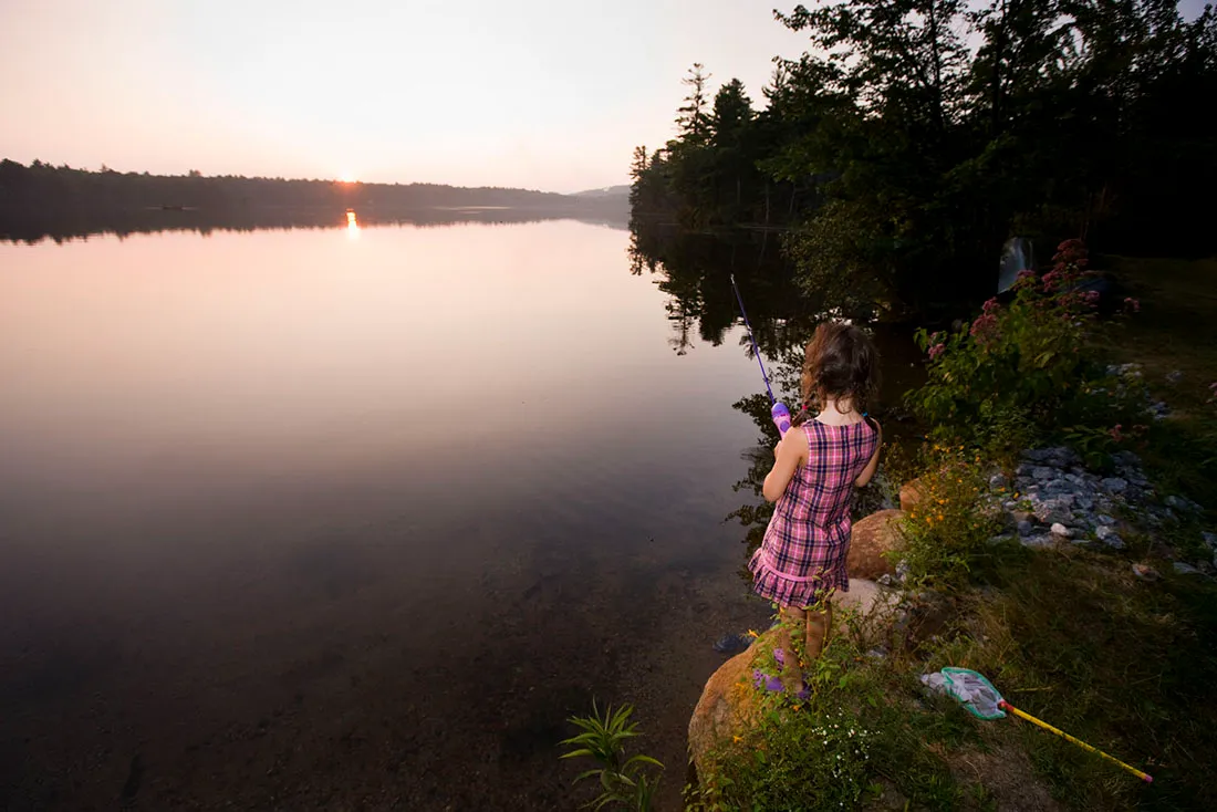 Girl fishing from the bank of a serene lake at sunset