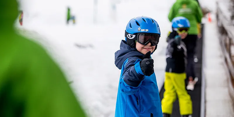 Kid on a magic carpet ski lift turning over his shoulder and giving a thumbs up