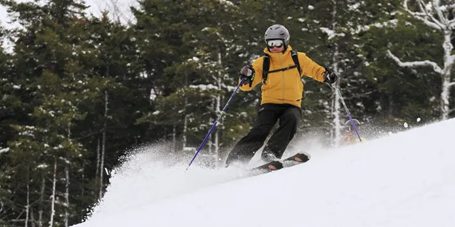 Skier in a yellow jacket kicking up powder on a turn