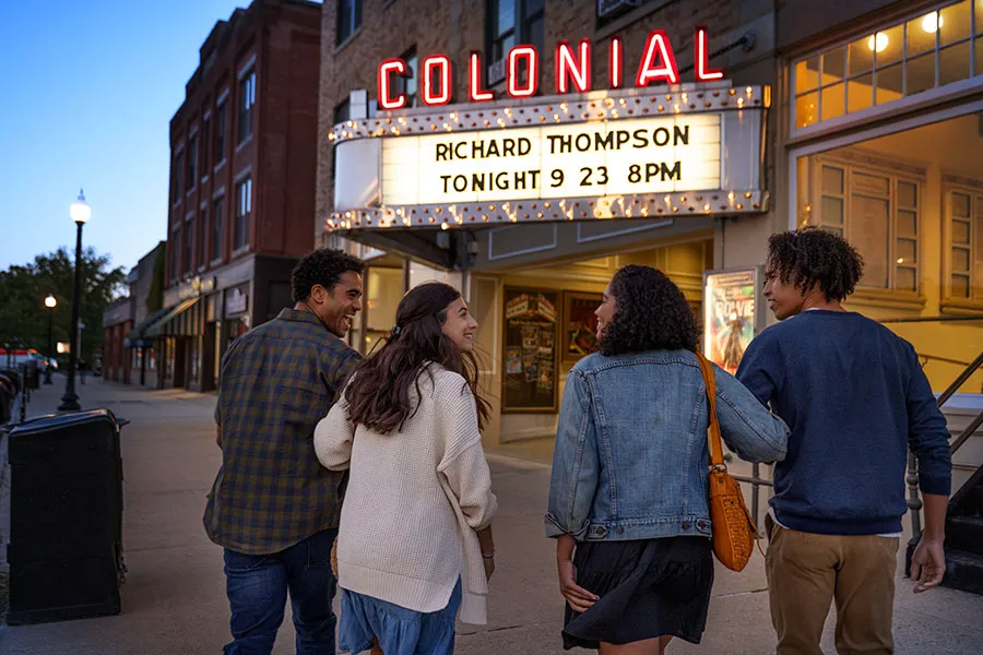 Two couples smiling, waling arm-in-arm into a theatre with a lighted marquee out front