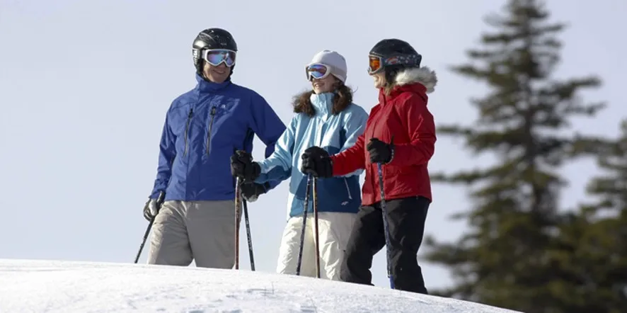 Three skiers in ski goggles and winter gear smiling at each other at the top of a run