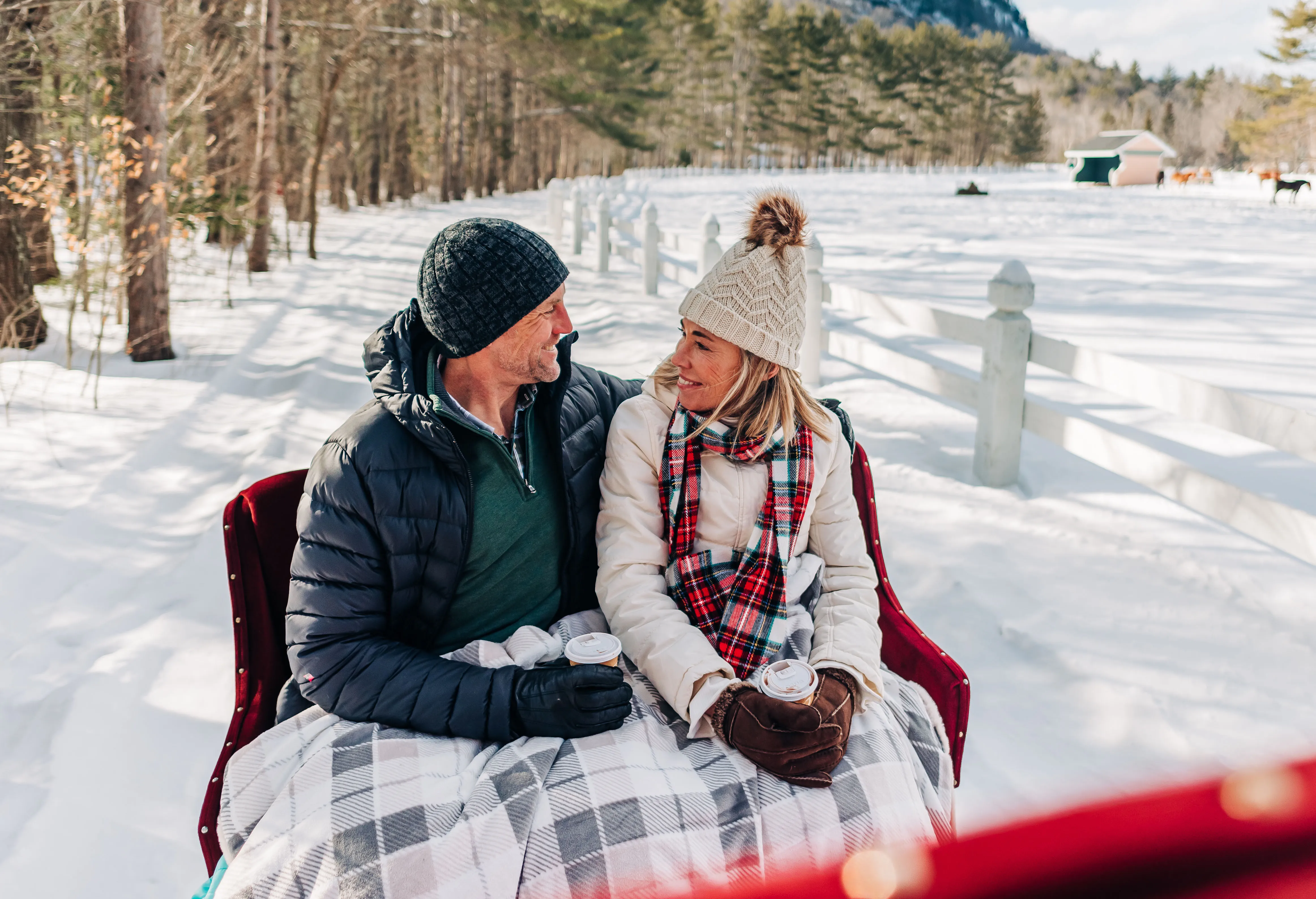 Couple enjoying a hot beverage on the back of a horse-drawn sled, their legs covered by a warm blanket