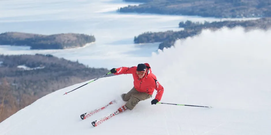 Skier in a red jacket kicking up powder on a turn, a frozen lake and evergreen trees in the valley in the background